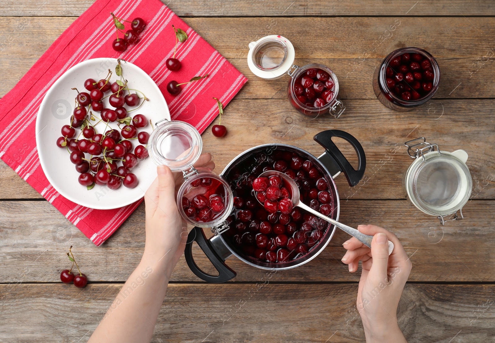 Photo of Woman putting delicious pickled cherries into jar at wooden table, top view