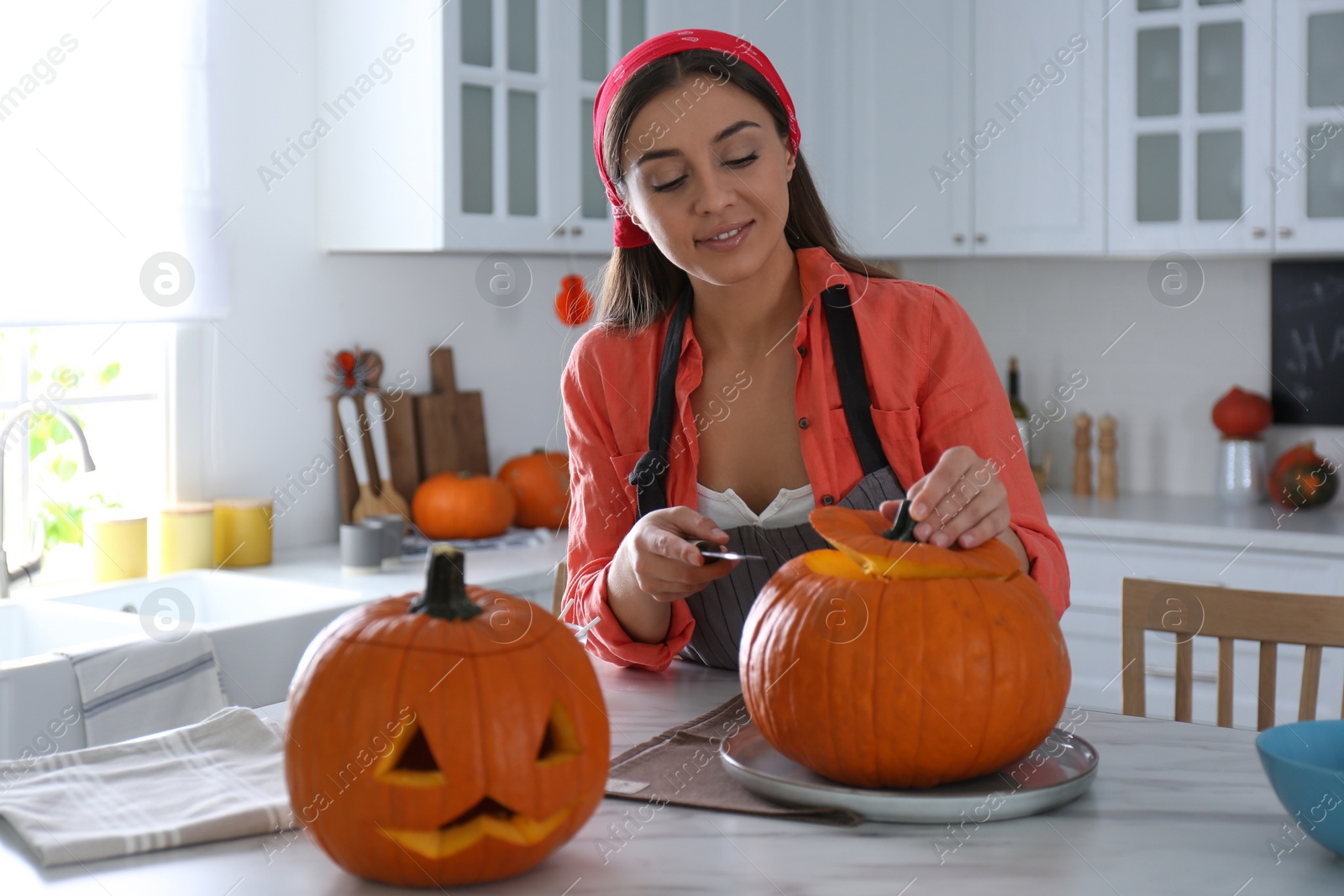 Photo of Woman making pumpkin jack o'lantern at table in kitchen. Halloween celebration