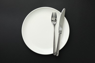 Photo of Clean plate, fork and knife on black table, top view