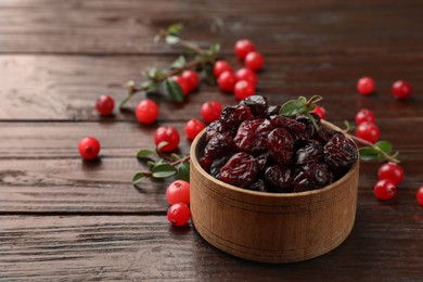 Photo of Tasty dried cranberries in bowl, fresh ones and leaves on wooden table, closeup. Space for text