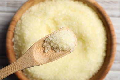 Spoon with yellow sea salt above bowl on wooden table, closeup. Top view