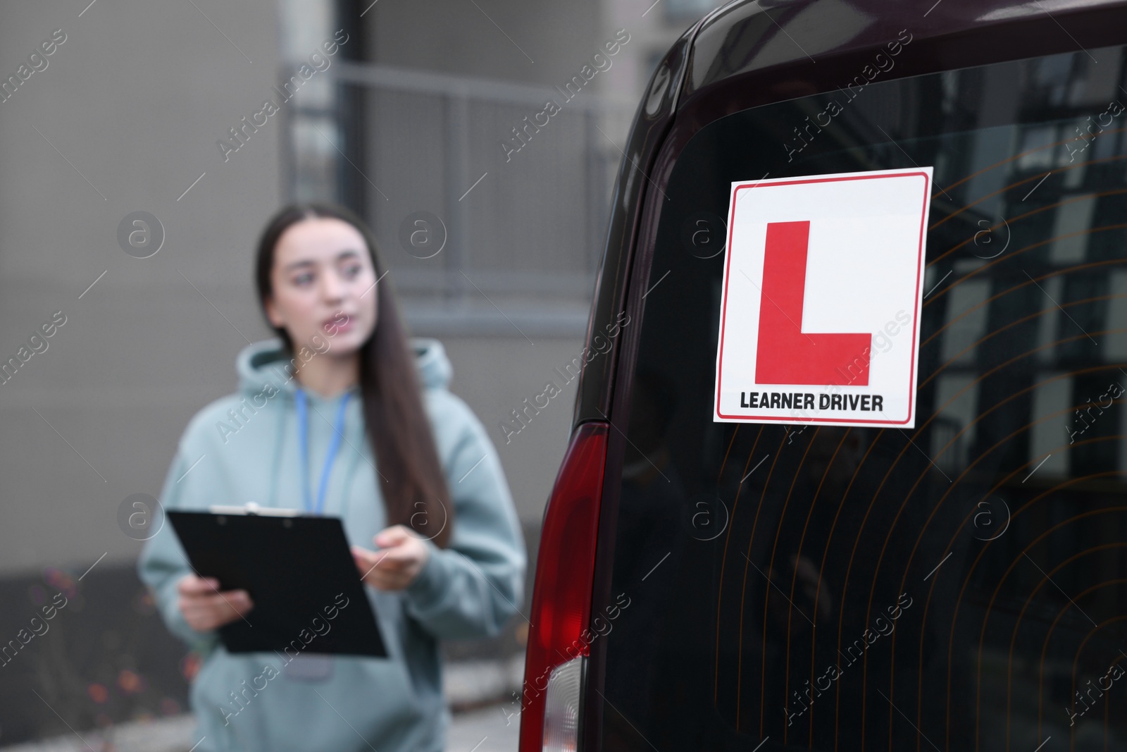 Photo of Instructor with clipboard near car outdoors, selective focus on L-plate. Driving school