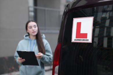 Instructor with clipboard near car outdoors, selective focus on L-plate. Driving school