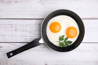 Tasty fried eggs with parsley in pan on white wooden table, top view