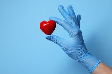 Doctor wearing medical glove holding decorative heart on light blue background, closeup