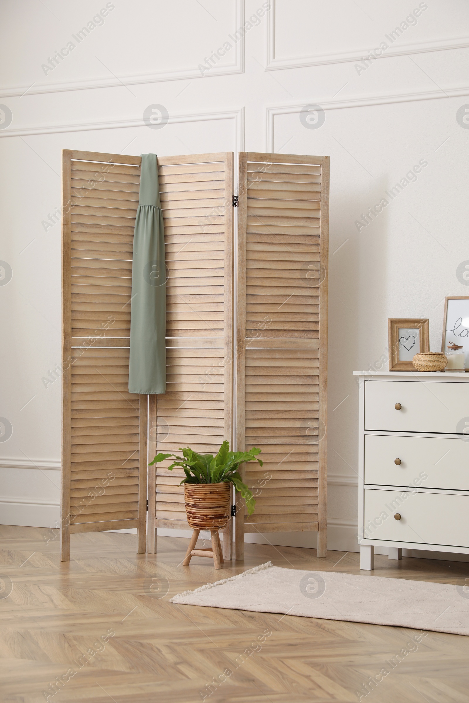 Photo of Stylish room with wooden folding screen and chest of drawers near white wall. Interior design