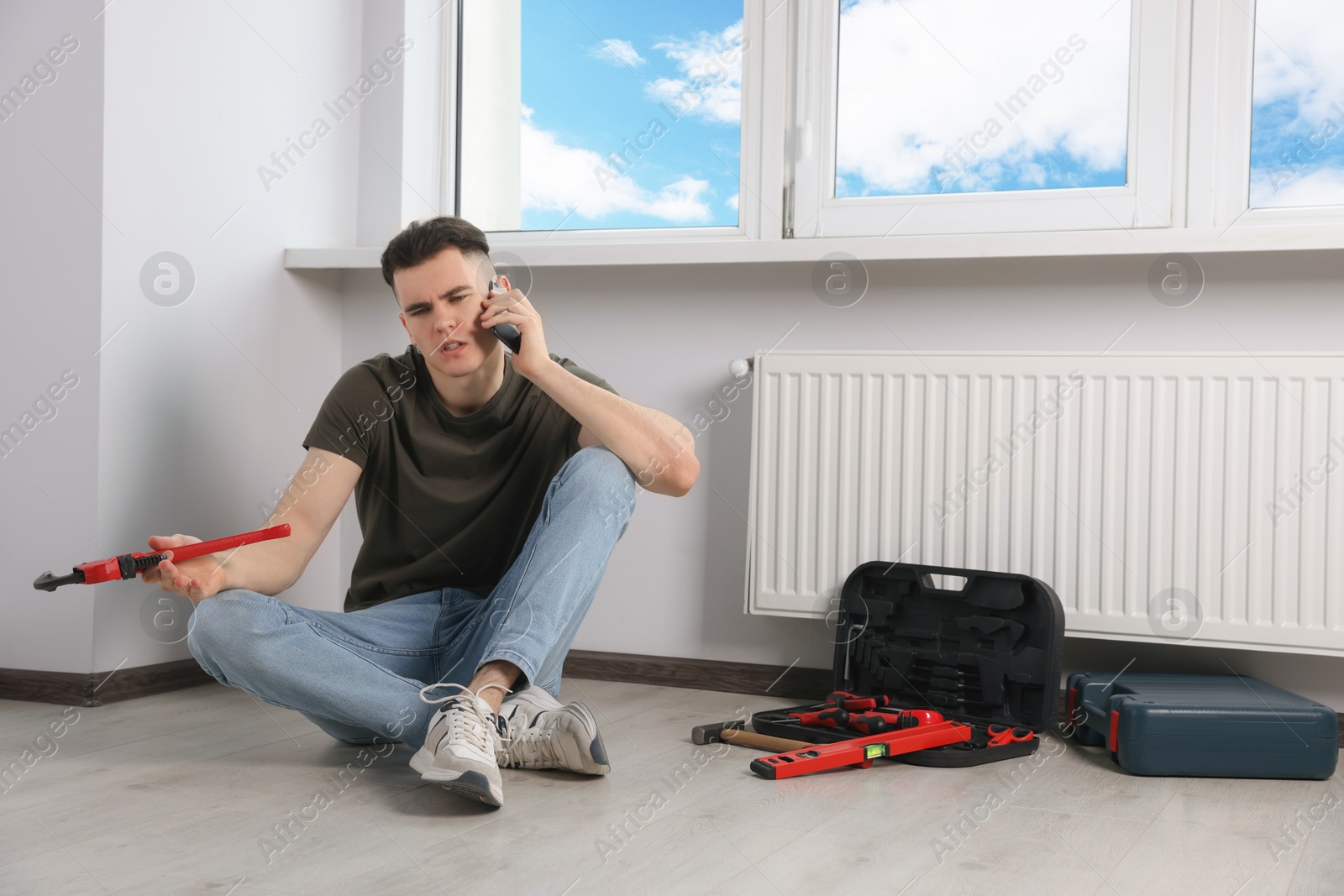 Photo of Handyman with pipe wrench talking on phone near radiator in room