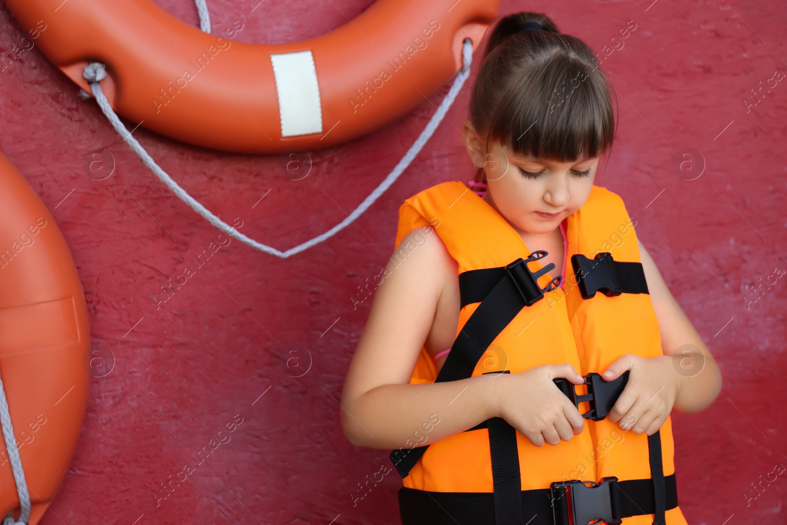 Photo of Little girl putting on orange life vest near red wall with safety rings