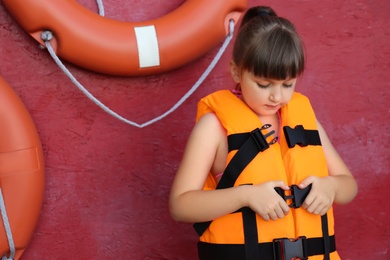 Little girl putting on orange life vest near red wall with safety rings