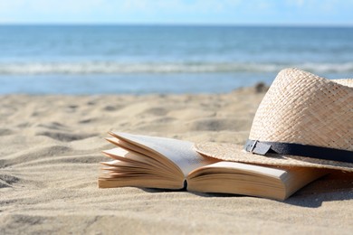 Photo of Open book and straw hat on sandy beach near sea, space for text