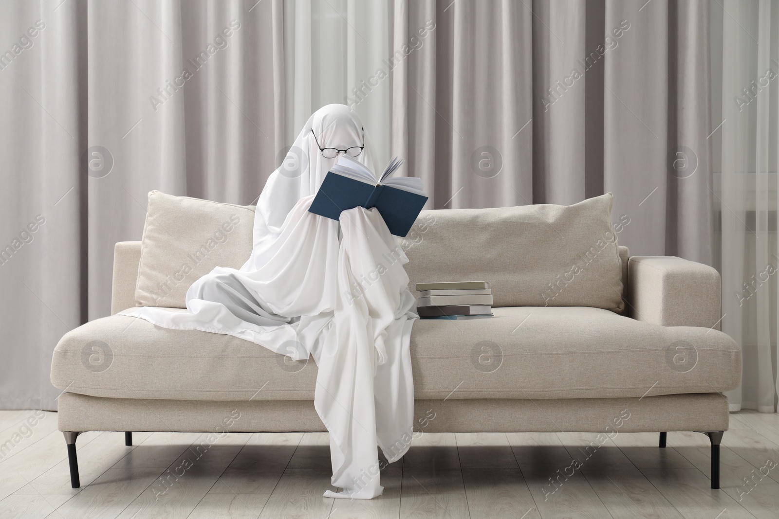 Photo of Creepy ghost. Person covered with white sheet reading book on sofa at home, space for text