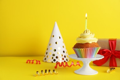 Photo of Delicious birthday cupcake with candle near gift box and party hat on yellow background, space for text