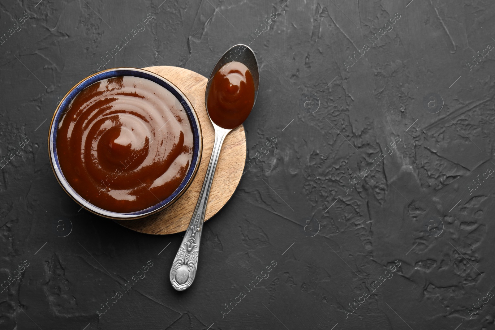 Photo of Tasty barbeque sauce in bowl and spoon on dark textured table, top view. Space for text