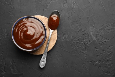 Tasty barbeque sauce in bowl and spoon on dark textured table, top view. Space for text