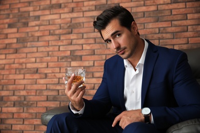 Young man with glass of whiskey near brick wall indoors