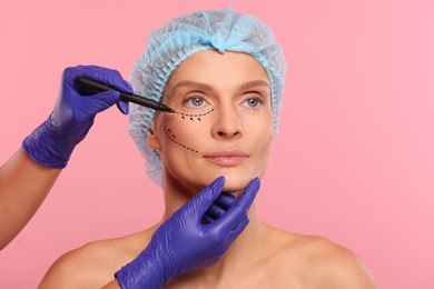 Image of Woman preparing for cosmetic surgery, pink background. Doctor drawing markings on her face, closeup