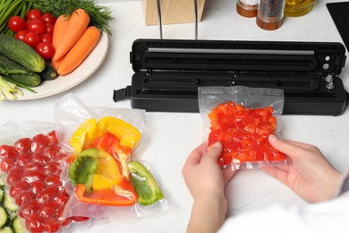 Photo of Woman using sealer for vacuum packing with plastic bag of red pepper at white table, closeup