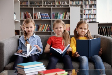 Photo of Happy little girls reading books on sofa in library