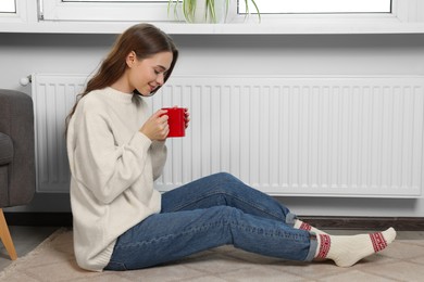 Woman holding cup with hot drink near heating radiator indoors