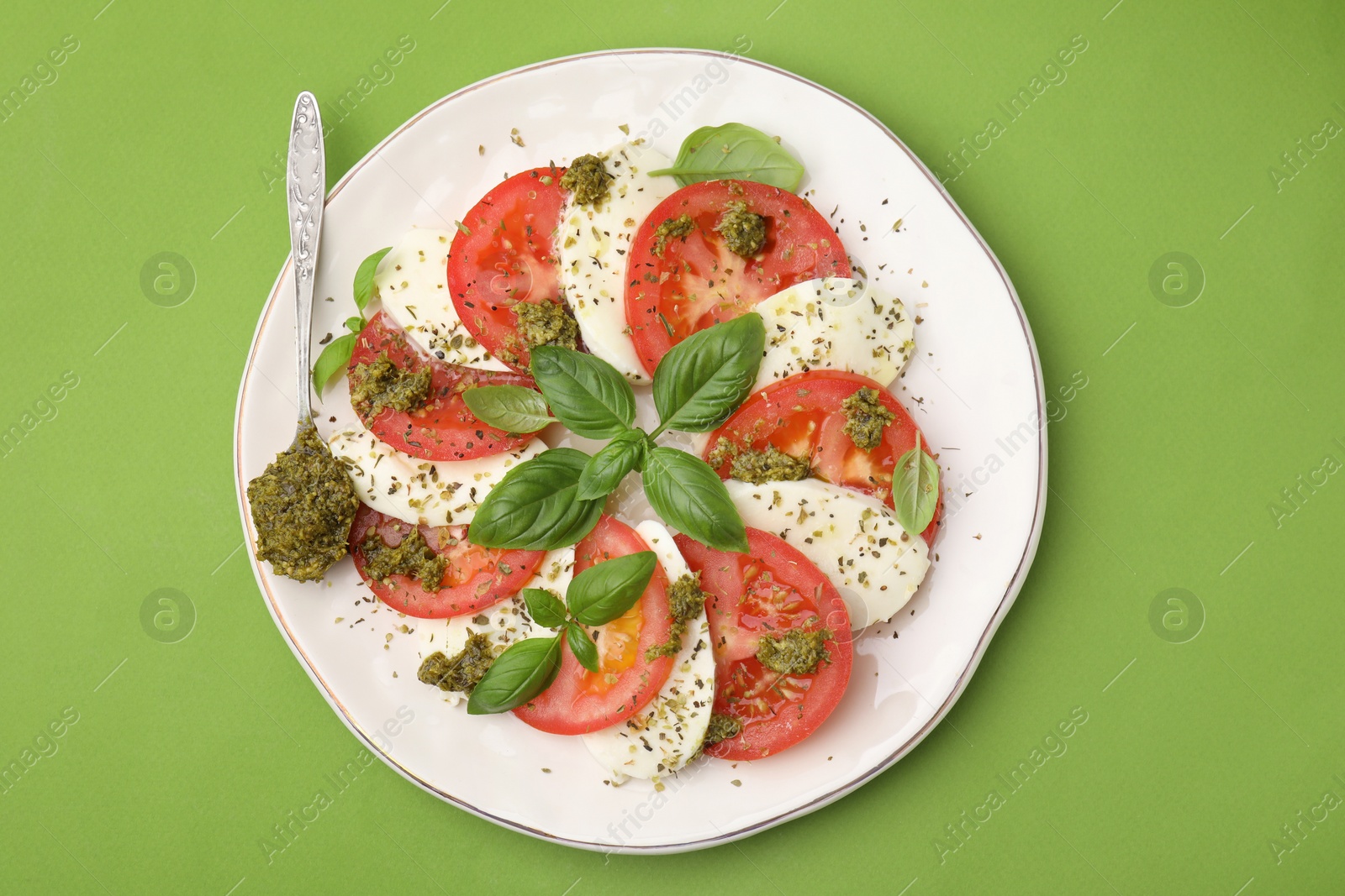 Photo of Plate of delicious Caprese salad and spoon with pesto sauce on green table, top view