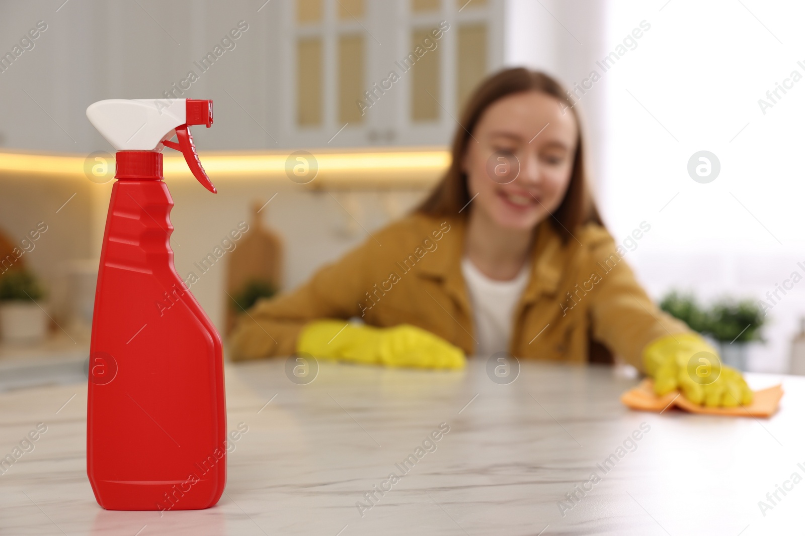 Photo of Woman with microfiber cloth cleaning white marble table in kitchen, focus on spray bottle