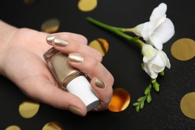 Woman with golden manicure holding nail polish bottle on black background, closeup