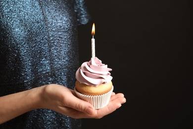 Photo of Woman holding delicious birthday cupcake with burning candle on black background, closeup