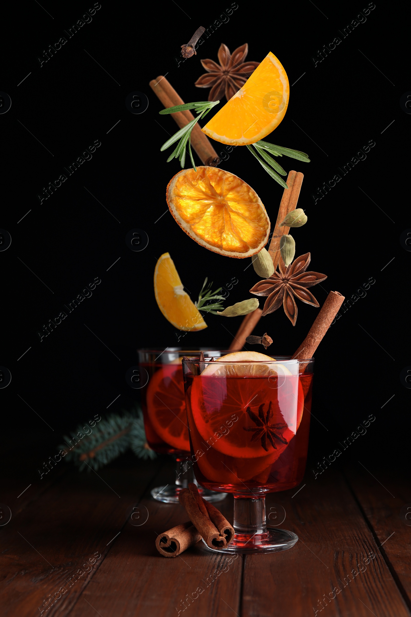 Image of Cut orange and different spices falling into glass cups of mulled wine on wooden table against black background