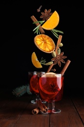 Cut orange and different spices falling into glass cups of mulled wine on wooden table against black background