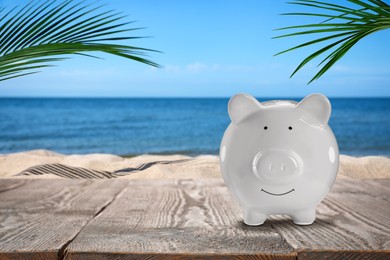 Image of Saving money for summer vacation. Piggy bank on wooden surface near sandy beach and sea, space for text