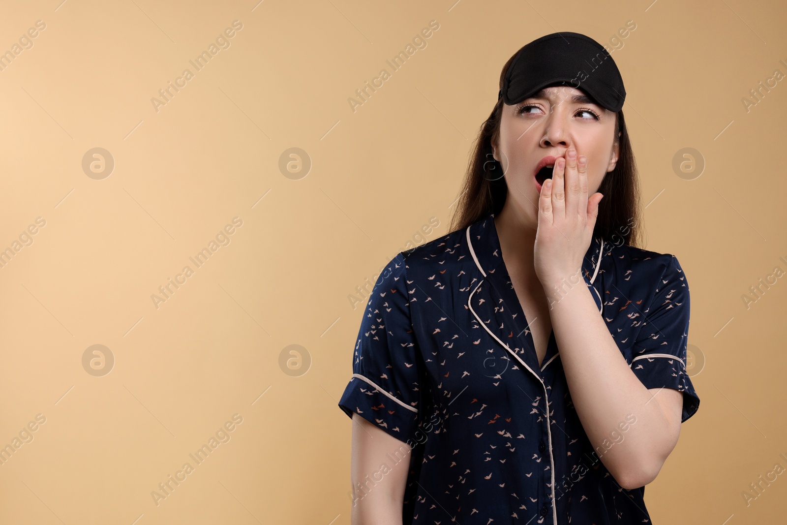 Photo of Tired young woman with sleep mask yawning on beige background, space for text. Insomnia problem