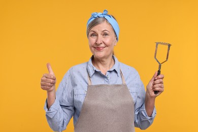 Photo of Happy housewife with potato masher showing thumbs up on orange background