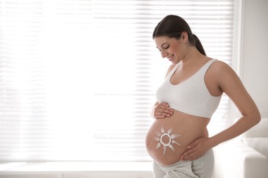 Young pregnant woman with sun protection cream on belly near window, space for text