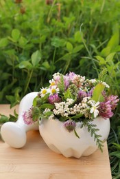 Photo of Ceramic mortar with pestle, different wildflowers and herbs on wooden board in meadow