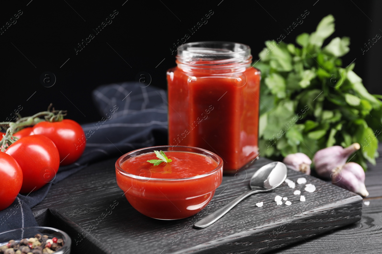 Photo of Delicious ketchup, spoon and spices on black wooden table. Tomato sauce