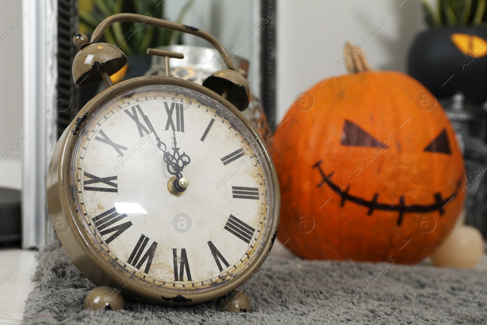 Photo of Old alarm clock and pumpkin with drawn spooky face on rug indoors. Halloween decor