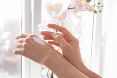 Photo of Young woman applying cream onto hands against window