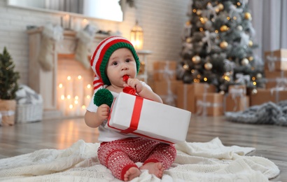 Cute little baby with elf hat and Christmas gift on floor at home