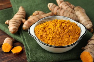 Bowl with aromatic turmeric powder and cut roots on wooden table, closeup