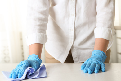 Photo of Woman in gloves wiping white table with rag indoors, closeup