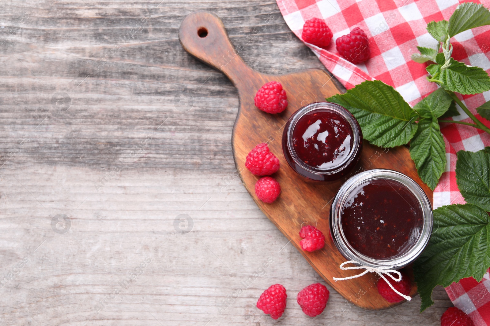 Photo of Delicious raspberry jam, fresh berries and green leaves on wooden table, flat lay. Space for text