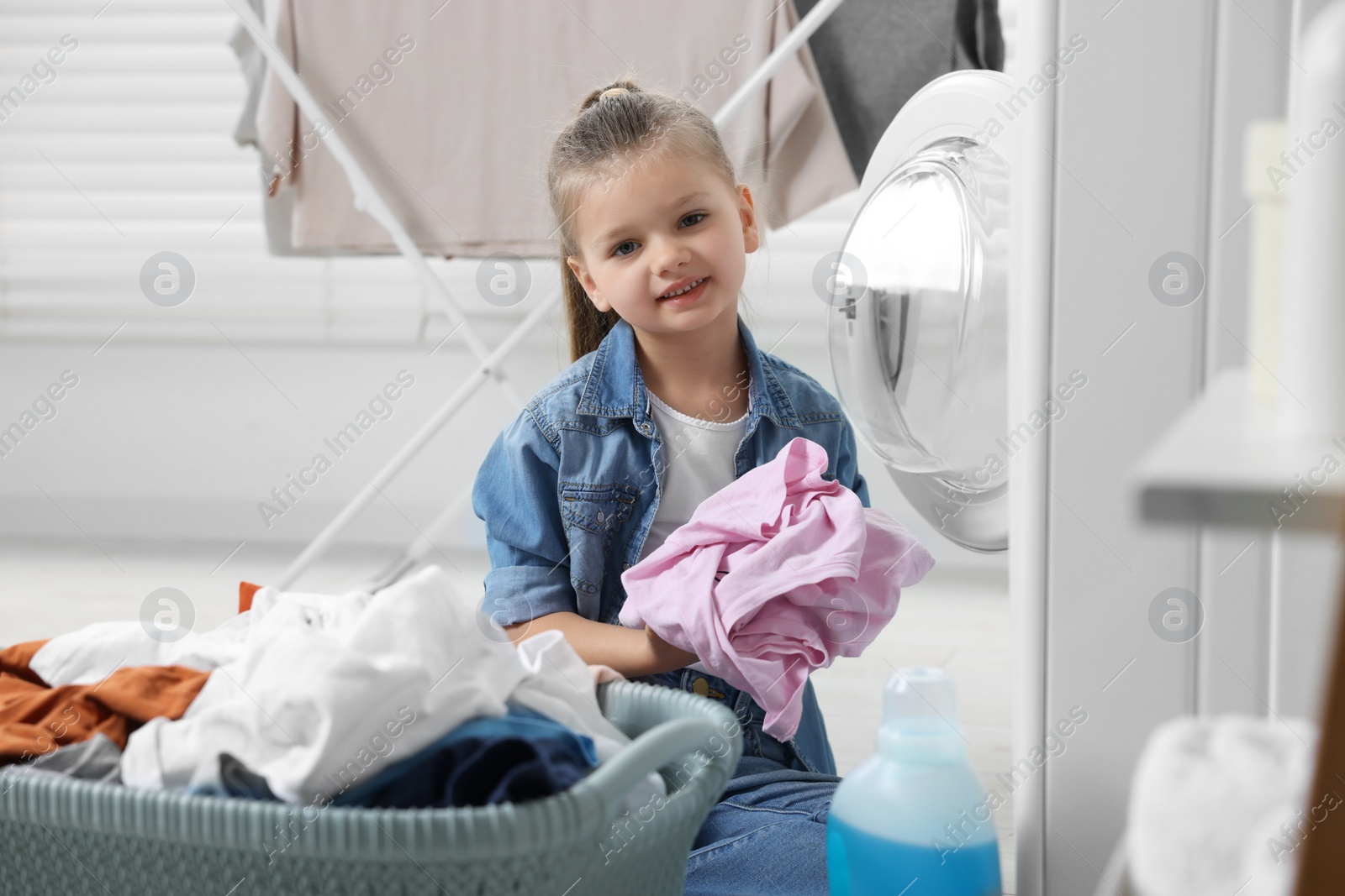 Photo of Little girl putting dirty clothes into washing machine in bathroom