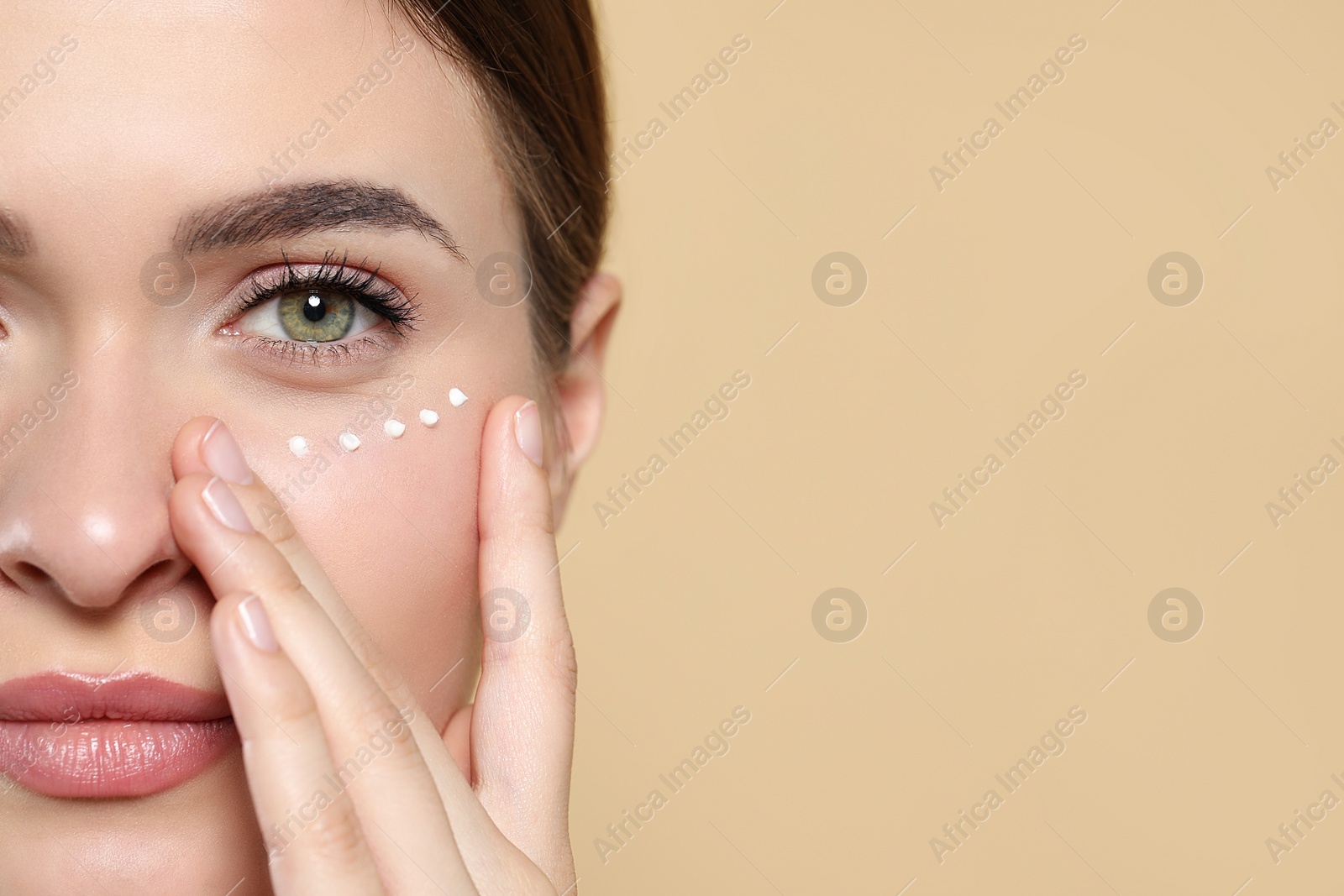 Photo of Young woman with cream around eye on beige background, closeup. Space for text