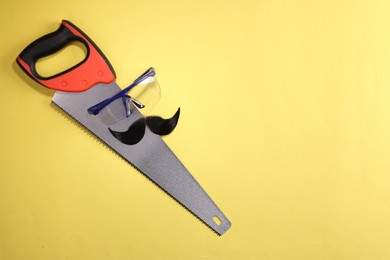 Man's face made of artificial mustache, safety glasses and hand saw on yellow background, top view. Space for text