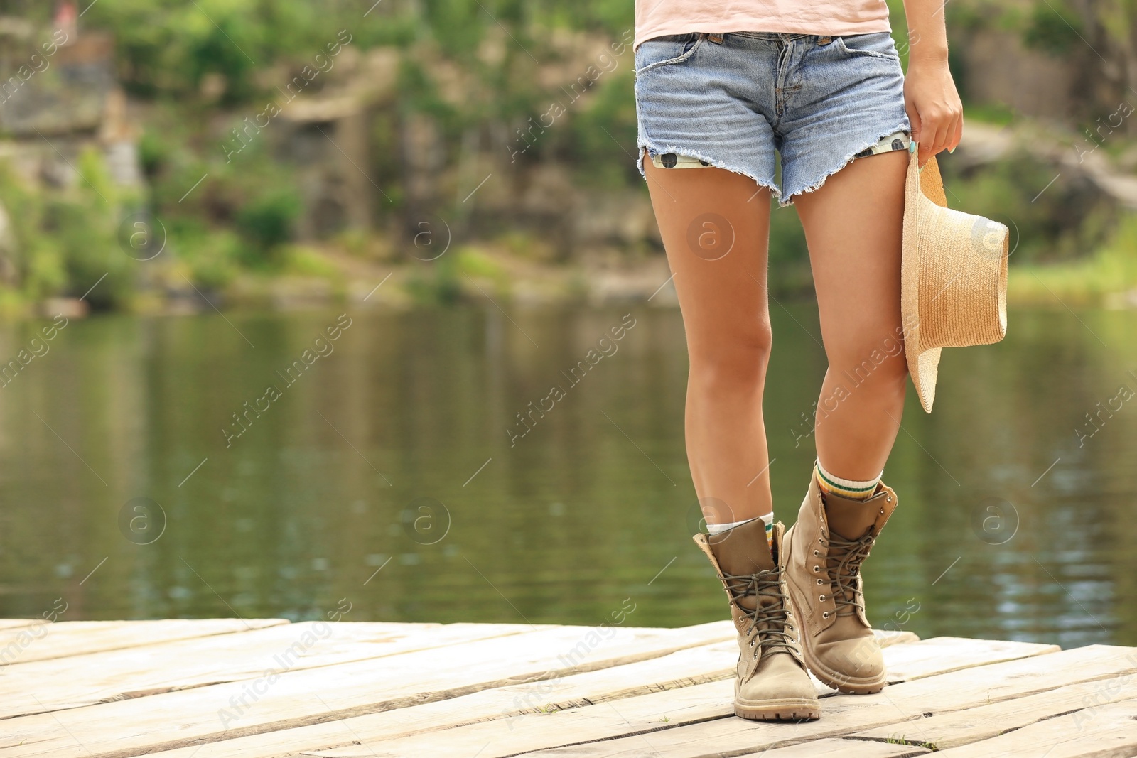 Photo of Young woman with hat on wooden pier near lake, focus on legs. Camping season
