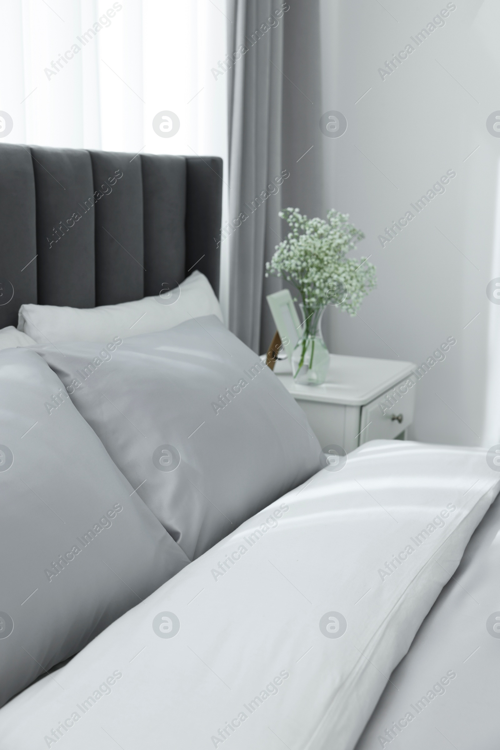 Photo of Comfortable bed and vase with beautiful flowers on white bedside table in room. Stylish interior