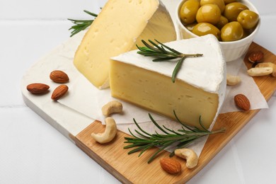 Photo of Pieces of tasty camembert cheese, rosemary, nuts and olives on white tiled table, closeup