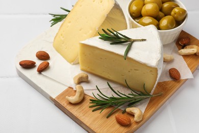 Pieces of tasty camembert cheese, rosemary, nuts and olives on white tiled table, closeup