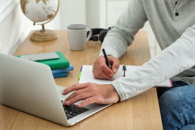 Photo of Man with laptop and notebook learning at wooden table indoors, closeup
