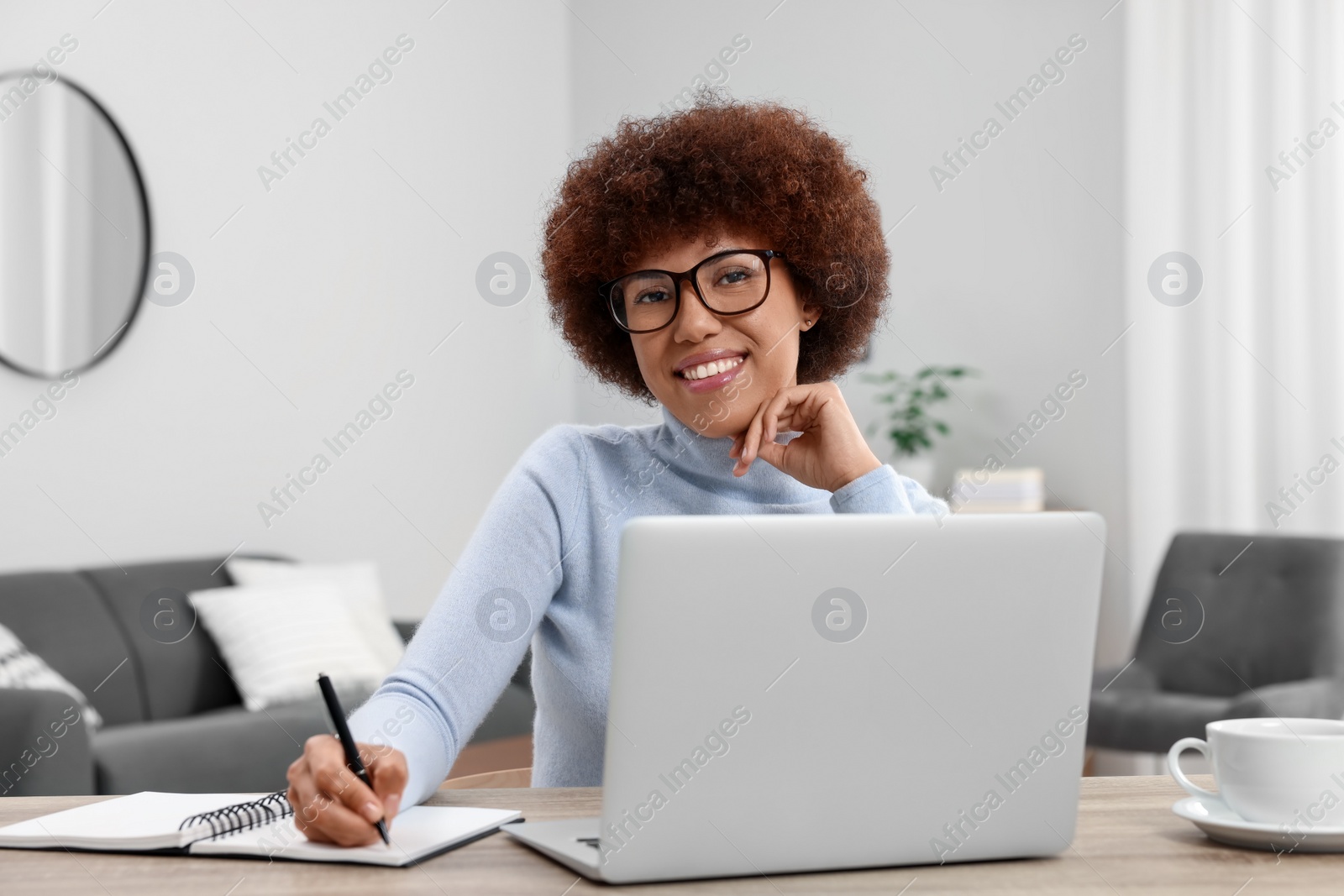 Photo of Beautiful young woman using laptop and writing in notebook at wooden desk in room